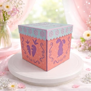Peut inclure: Une boîte décorative sur le thème de Pâques, avec un extérieur rose clair, ornée de motifs découpés de carottes et de lapins. La boîte a une bordure festonnée bleu clair et un couvercle assorti. La boîte est posée sur un présentoir à gâteau blanc.