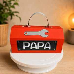 Puede incluir: Una caja de herramientas roja con asa plateada y un dibujo de llave inglesa plateada. La palabra "PAPA" está escrita en blanco sobre un rectángulo negro. La caja de herramientas está sobre un soporte blanco para tartas.