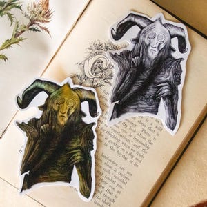 Puede incluir: Dos pegatinas que representan una criatura fantástica con cuernos, una en verde y marrón, la otra en blanco y negro. Se colocan sobre un libro abierto con páginas amarillentas, evocando una estética vintage.