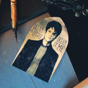 May include: Illustration of a person with the text "Dream a Little Dream of Me". The artwork is in black and white, with the person wearing a dark jacket. The image is on a light tan background.