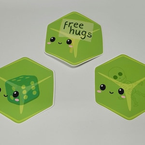 May include: Three green, cube-shaped stickers with cartoon faces. One sticker has a die inside, another has a skeleton, and the third has a sign that says "free hugs". Each sticker has a white border.