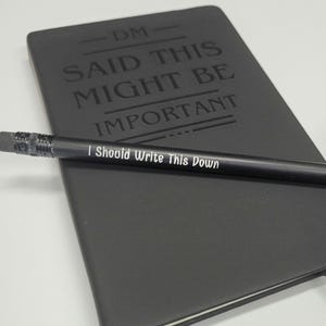 May include: A black notebook with the text "DM SAID THIS MIGHT BE IMPORTANT I Should Write This Down" on the front cover. A black pencil is lying on top of the notebook.