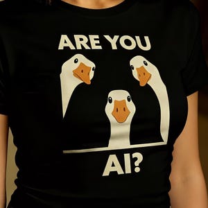 Op de afbeelding: Zwart T-shirt met de tekst "ARE YOU AI?" en een afbeelding van drie witte ganzen met oranje snavels. Het T-shirt is gemaakt van een zacht materiaal en is geschikt voor casual wear.