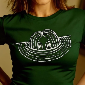 Op de afbeelding: Donkergroen t-shirt met een witte lijntekening van een gezicht in een cirkelvormig ontwerp. Het shirt is effen van kleur en heeft korte mouwen. Het ontwerp is gecentreerd op de voorkant van het shirt.