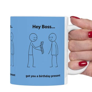 Puede incluir: Taza de cerámica blanca con un diseño de dibujos animados humorístico. La taza presenta figuras de palo y el texto "Hey Boss... got you a birthday present" sobre un fondo azul claro. La taza está sostenida por una mano con esmalte de uñas rojo.