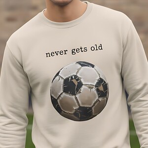 Op de afbeelding: Beige sweatshirt met een voetbalafbeelding en de tekst "never gets old". De voetbal is zwart en wit. De sweatshirt heeft een ronde hals.