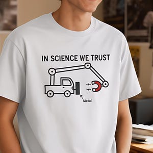 Op de afbeelding: Wit t-shirt met de tekst "IN SCIENCE WE TRUST" en een afbeelding van een vrachtwagen met een magnetische arm die metaal optilt. De afbeelding is in zwart en rood. De persoon draagt een blauwe spijkerbroek.