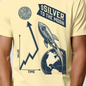 以下が含まれることがあります： グラフィックデザインの薄い黄色のTシャツ。デザインには、コイン、上昇傾向のグラフ、ロケット、テキスト「$SILVER TO THE MOON」が含まれています。ロケットは地球から打ち上げられています。