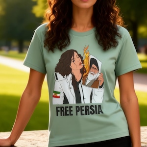 Peut inclure: Un t-shirt vert clair avec le texte "FREE PERSIA" et un graphisme représentant une femme et un homme. La femme porte une veste blanche avec le drapeau iranien. La personne se tient devant un mur en pierre.