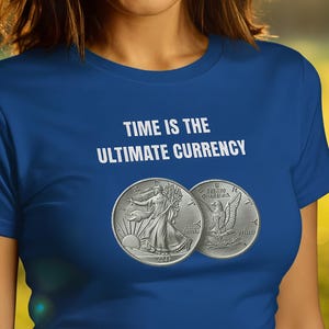 May include: A woman wearing a blue t-shirt with the text "TIME IS THE ULTIMATE CURRENCY" in white. Two silver coins are printed on the shirt. The woman is smiling in front of a blurred green background.