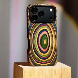 Op de afbeelding: Een houten telefoonhoesje met een kleurrijk, concentrisch cirkelontwerp. De hoes heeft een zwarte cameramodule en wordt gepresenteerd op een klein houten blok. Het ontwerp bevat ringen van groen, geel, blauw en rood.