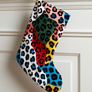 May include: A colorful Christmas stocking with a patchwork design. The stocking features various leopard print patterns in white, black, red, blue, green, and yellow. It hangs from a gold hook with a burgundy ribbon, against a white door.