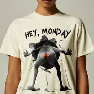 Op de afbeelding: Crèmekleurig T-shirt met de zwarte tekst "HEY, MONDAY". Het shirt heeft een afbeelding van een figuur die door een muur breekt, met een rood lippenontwerp. Het T-shirt is een casual kledingstuk.