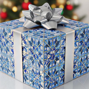 Op de afbeelding: Een vierkante geschenkdoos met een blauw en wit glas-in-loodpatroon, zilveren lint en een grote zilveren strik. De doos staat op een wit oppervlak met een vage kerstboom op de achtergrond.