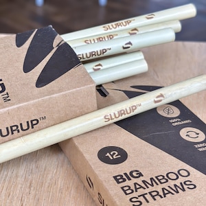 Può includere: Primo piano di cannucce di bambù e la loro confezione. Le cannucce sono beige chiaro con la scritta "SLURUP" stampata sopra. La confezione è marrone con le scritte "SLURUP" e "BIG BAMBOO STRAWS" stampate. La confezione ha anche un "12" stampato.