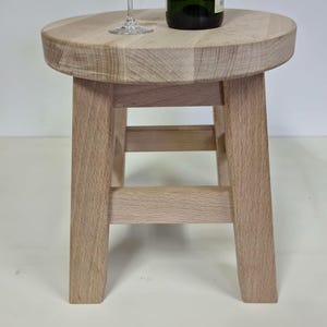 Könnte beinhalten: Ein kleiner, runder Holzschemel mit einer Champagnerflasche und einem Glas Champagner. Der Hocker ist aus hellem Holz gefertigt und hat vier Beine. Die Flasche ist grün mit einem goldenen Deckel. Das Glas ist klar.