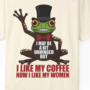 Könnte beinhalten: Cremefarbenes T-Shirt mit einer Cartoon-Froschfigur, die einen Zylinder und eine Fliege trägt. Der Frosch hat große orangefarbene Augen und ein grünes Gesicht. Der Text auf dem Shirt lautet "I MAY BE A BIT UNHINGED BUT I LIKE MY COFFEE HOW I LIKE MY WOMEN."