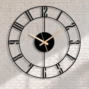 Könnte beinhalten: Eine große Wanduhr aus schwarzem Metall mit römischen Ziffern und einem kreisförmigen Design. Die Uhrzeiger sind hellbraun. Die Uhr ist an einer weißen Backsteinmauer befestigt. Die Uhr ist ein dekoratives Element in einem Wohnzimmer.