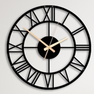 May include: A large, circular wall clock featuring a black metal frame with open-work Roman numerals. The clock has a solid black centre disc and thin, light-coloured metallic hands that contrast against the dark structure of the clock face.