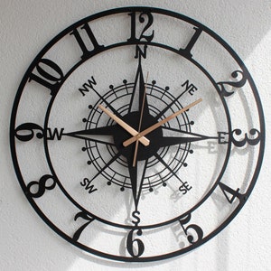Könnte beinhalten: Schwarze Wanduhr aus Metall mit Kompassdesign. Das Zifferblatt der Uhr zeigt römische Ziffern und eine Kompassrose mit den Richtungen Nord, Süd, Ost und West.