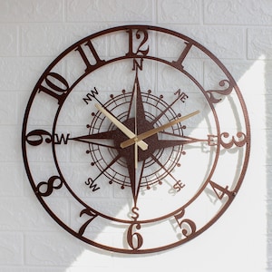 May include: A round wall clock featuring a dark brown metal frame with cut-out Arabic numerals from 1 to 12. The center displays a detailed compass rose design with cardinal directions labeled N, S, E, W, NW, NE, SW, and SE. Two gold-toned hands.