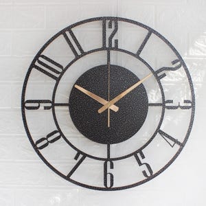 May include: A round, black metal wall clock with a textured centre and gold-coloured hands. The clock face features Roman numerals and a minimalist design, suitable for home decor. The clock's diameter is approximately 30 cm.