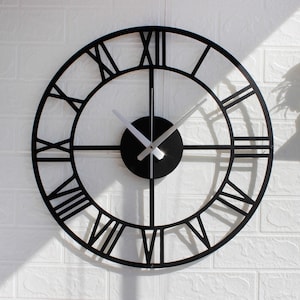 Może przedstawiać: Czarny, otwarty zegar ścienny z rzymskimi cyframi. Zegar ma okrągły kształt z czarnym środkiem i srebrnymi wskazówkami. Zegar jest zamontowany na białej ceglanej ścianie.