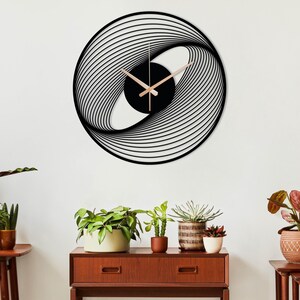May include: A modern, black and gold wall clock with a unique, abstract design. The clock features a black circular center with gold-colored hands and a swirling, layered pattern around the perimeter. The clock is mounted on a white wall.