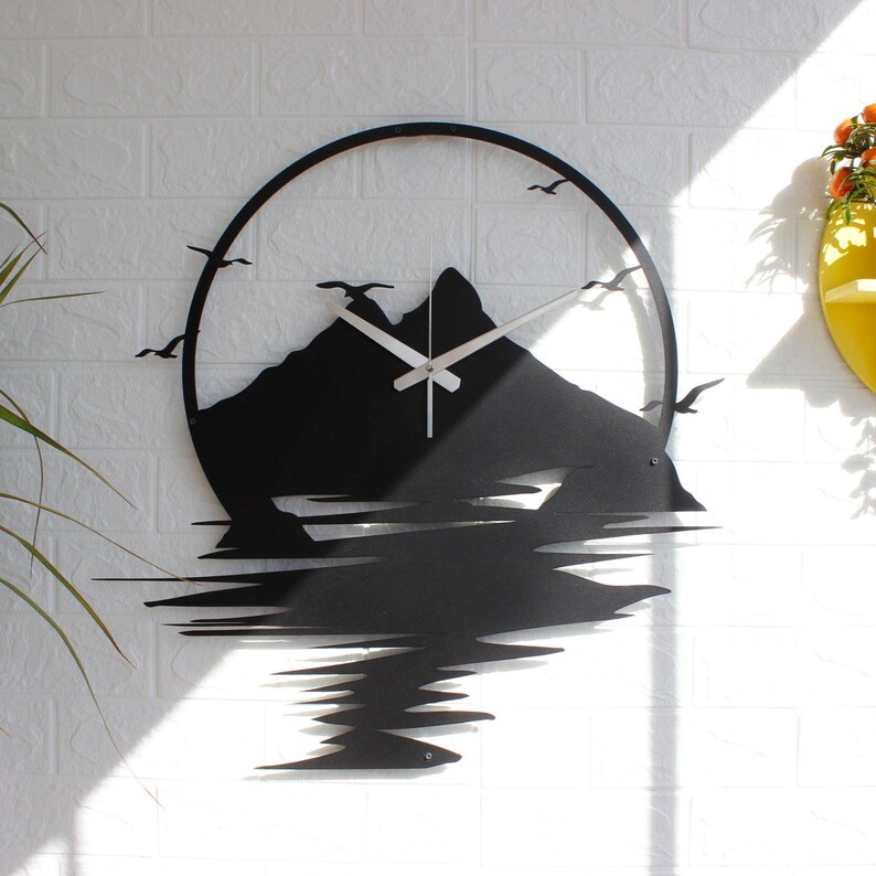 May include: A circular black metal wall clock featuring a silhouette of a mountain range and flying birds. The design includes a reflection of the mountains at the bottom, with silver-colored clock hands positioned in the center of the frame.
