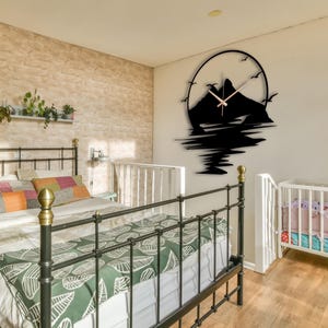 May include: A black circular wall clock featuring a silhouette of a mountain range and birds in flight. The clock face has thin, light-colored hands centered over the mountain design, which extends downward with a reflection-like pattern.