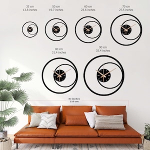 Pu&ograve; includere: Una collezione di orologi da parete neri moderni di varie dimensioni, da 35 cm a 90 cm di diametro. Ogni orologio presenta un quadrante nero con lancette chiare e un design unico a doppio cerchio. Gli orologi sono esposti su una parete bianca sopra un divano in pelle marrone.