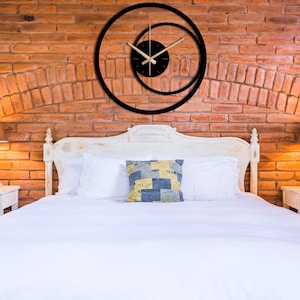 Pu&ograve; includere: Orologio da parete moderno nero con un design circolare unico. L'orologio ha un quadrante nero con lancette beige e una lancetta dei secondi. L'orologio &egrave; montato su un muro di mattoni rossi sopra un letto bianco.