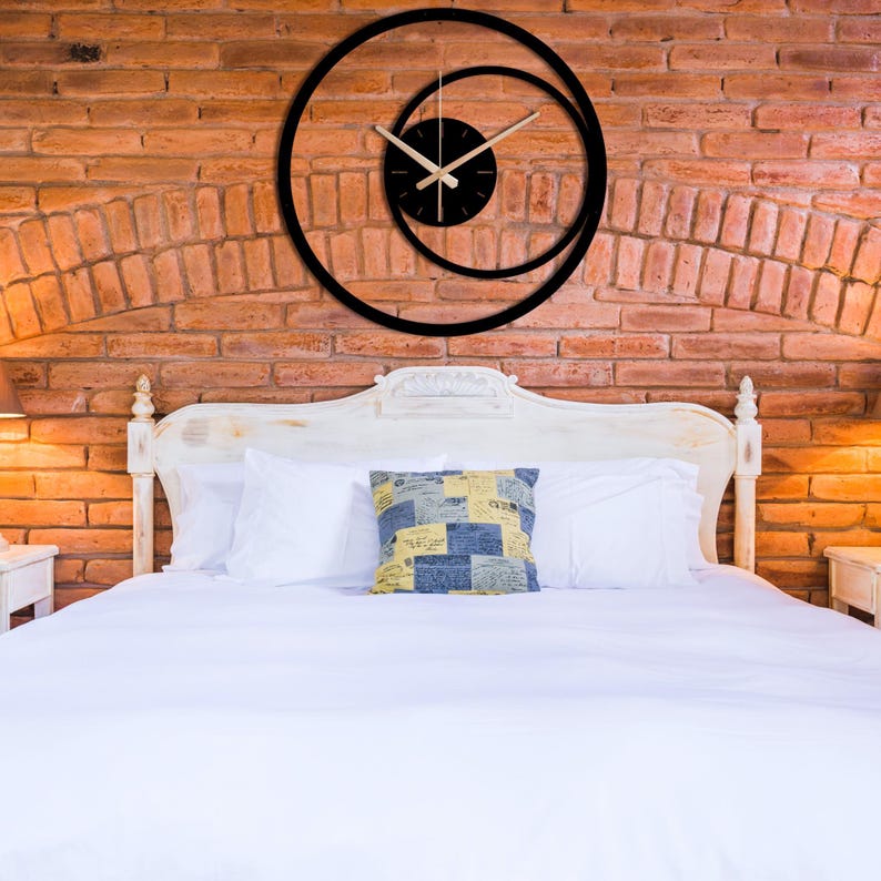 Pu&ograve; includere: Orologio da parete moderno nero con un design circolare unico. L'orologio ha un quadrante nero con lancette beige e una lancetta dei secondi. L'orologio &egrave; montato su un muro di mattoni rossi sopra un letto bianco.