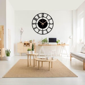 Op de afbeelding: Een zwarte metalen wandklok met Romeinse cijfers en een zwarte achtergrond. De klok hangt aan een witte muur in een woonkamer.