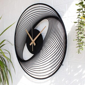 May include: Black and gold modern wall clock with an oval, layered design. The clock face is black with gold hands. The clock is mounted on a white brick wall. The clock's dimensions are approximately 20 inches in height and 15 inches in width.