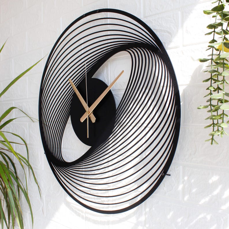 May include: Black and gold modern wall clock with an oval, layered design. The clock face is black with gold hands. The clock is mounted on a white brick wall. The clock's dimensions are approximately 20 inches in height and 15 inches in width.