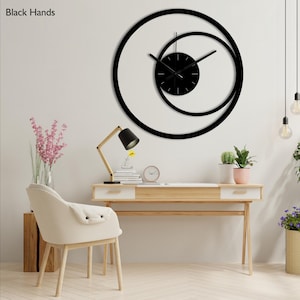 Pu&ograve; includere: Un moderno orologio da parete nero dal design unico. L'orologio presenta una cornice circolare con un cerchio pi&ugrave; piccolo e sovrapposto e un quadrante nero. Le lancette sono nere e l'orologio &egrave; montato su una parete bianca.