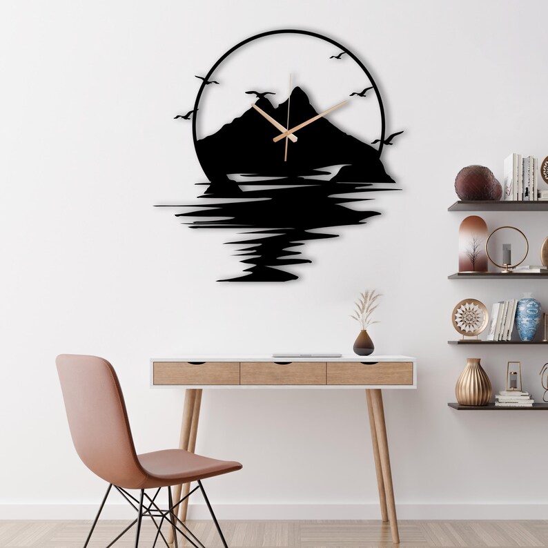 May include: A circular wall clock featuring a black silhouette of a mountain range and flying birds within a circular frame. The clock has thin, light-colored hands centered on the mountain design, with a black reflection effect extending below.