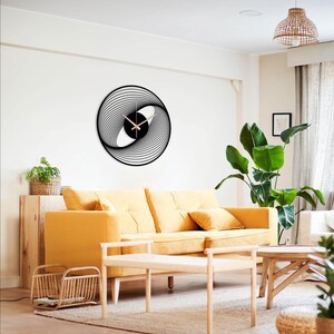 May include: A black and white wall clock with a unique spiral design. The clock features a black face with light-colored hands and a modern, abstract aesthetic. The clock is mounted on a white wall above a yellow sofa.