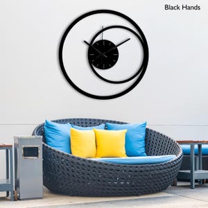 Pu&ograve; includere: Un moderno orologio da parete nero con un design unico caratterizzato da due forme circolari sovrapposte. Il quadrante dell'orologio &egrave; nero con lancette nere e il testo "Black Hands" &egrave; visibile. L'orologio &egrave; montato su una parete bianca.
