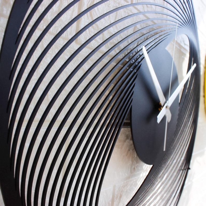 May include: Black, modern wall clock with a unique, swirling design. The clock face is a solid black circle with white hands. The clock's frame features a series of curved, black lines that create a spiral effect. The clock is a decorative piece.