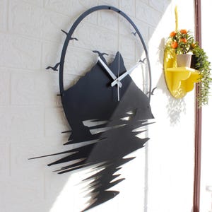 May include: A black metal wall clock featuring a circular frame and a mountain silhouette design. The clock face includes silver-colored hands and cut-out shapes of flying birds. The lower portion of the mountain design extends downward in jagged, abstract lines that mimic a reflection.