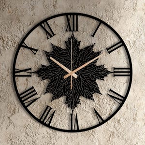 Puede incluir: Reloj de pared de metal negro con números romanos y un diseño de hoja de arce en el centro. Las manecillas del reloj son de color marrón claro. El reloj está montado en una pared texturizada de color claro.