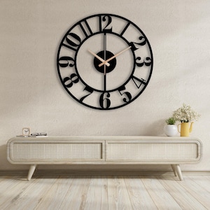 May include: A large circular wall clock featuring a black metal frame with cut-out numerals from one to twelve. The clock has a central black circular hub and two thin, light-coloured hands. The design is open, revealing the wall surface behind it.