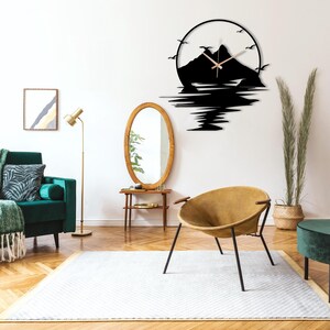 May include: A circular wall clock featuring a black silhouette of a mountain range and water reflection. The design includes flying birds and thin, light-colored clock hands centered within the mountain peak. The clock is mounted on a plain wall.