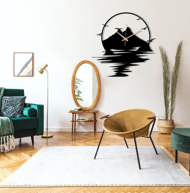 May include: A circular wall clock featuring a black silhouette of a mountain range and water reflection. The design includes flying birds and thin, light-colored clock hands centered within the mountain peak. The clock is mounted on a plain wall.