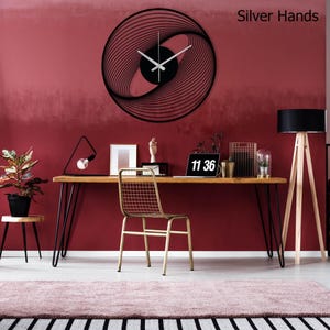 May include: A black, spiral-design wall clock with silver hands is mounted on a red wall. The clock's design features concentric lines that create a visual vortex. The time displayed on the clock is 11:36. The words "Silver Hands" are in the top right corner.
