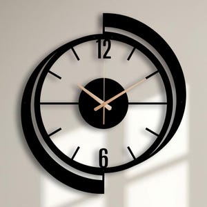 Può includere: Un orologio da parete circolare con una cornice nera e un anello esterno sfalsato a forma di mezzaluna. Il quadrante è trasparente con indicatori delle ore neri, i numeri 12 e 6 in grassetto, un cerchio centrale nero e lancette oro rosa.