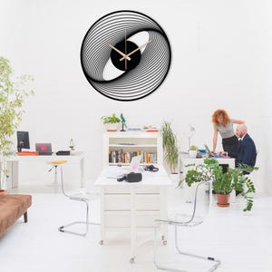 Modern spiral metal wall clock with black gold hands, hanging on a white wall in a workplace environment.