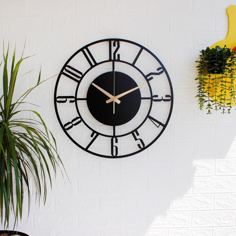 A large, modern, silent metal wall clock with a large green leafed flower pot on the left side and a yellow wall decoration on the right side, hanging on the wall on a white wall, black in color, with gold hands, and with Latin numerals.